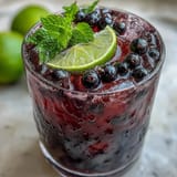 Vibrant purple Black Currant Mojito topped with soda bubbles, mint, and fresh black currants on a summer patio table.