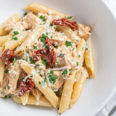 A close-up of Creamy Sun-Dried Tomato Chicken Pasta, highlighting the glossy, velvety sauce clinging to penne, chicken, and sliced sun-dried tomatoes, garnished with fresh basil.  